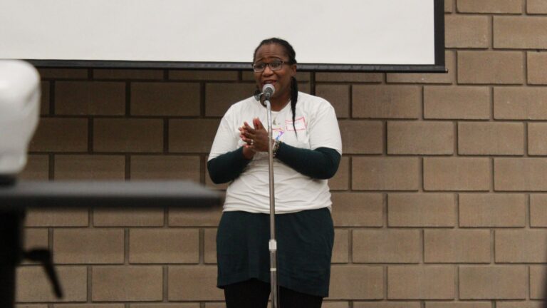 Woman in an "I AM" ToLSO shirt clapping at a microphone.