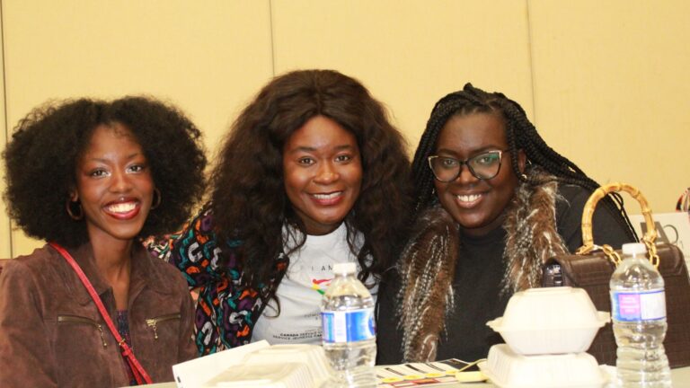 Three women seated together, the one in the center wearing an "I AM ToLSO" shirt.