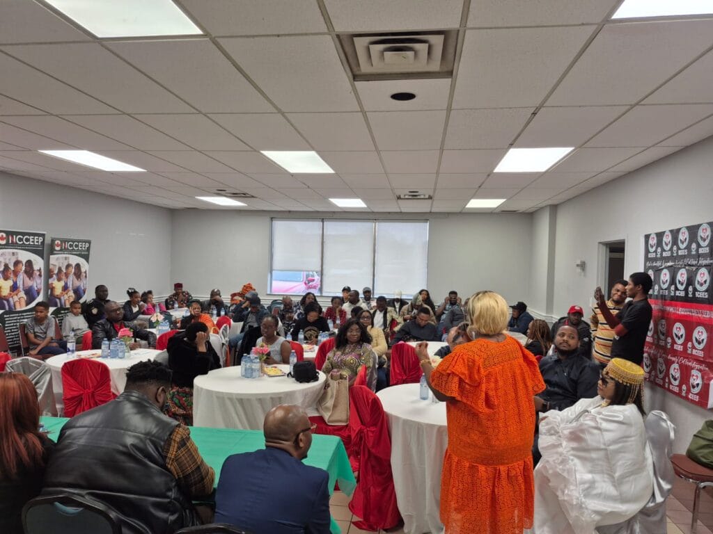 A wide view of the full hall, guests seated at round tables while a woman in orange stands to speak