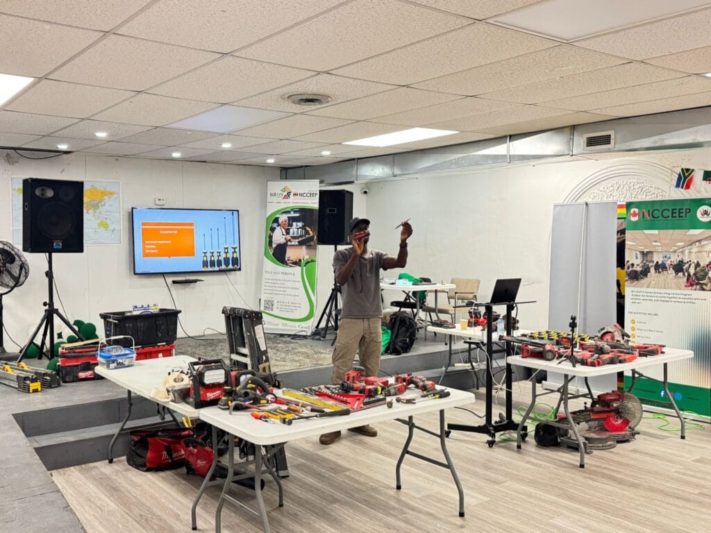 The workshop leader standing beside the screen with an arm raised, two long tables of power tools set out in front of him