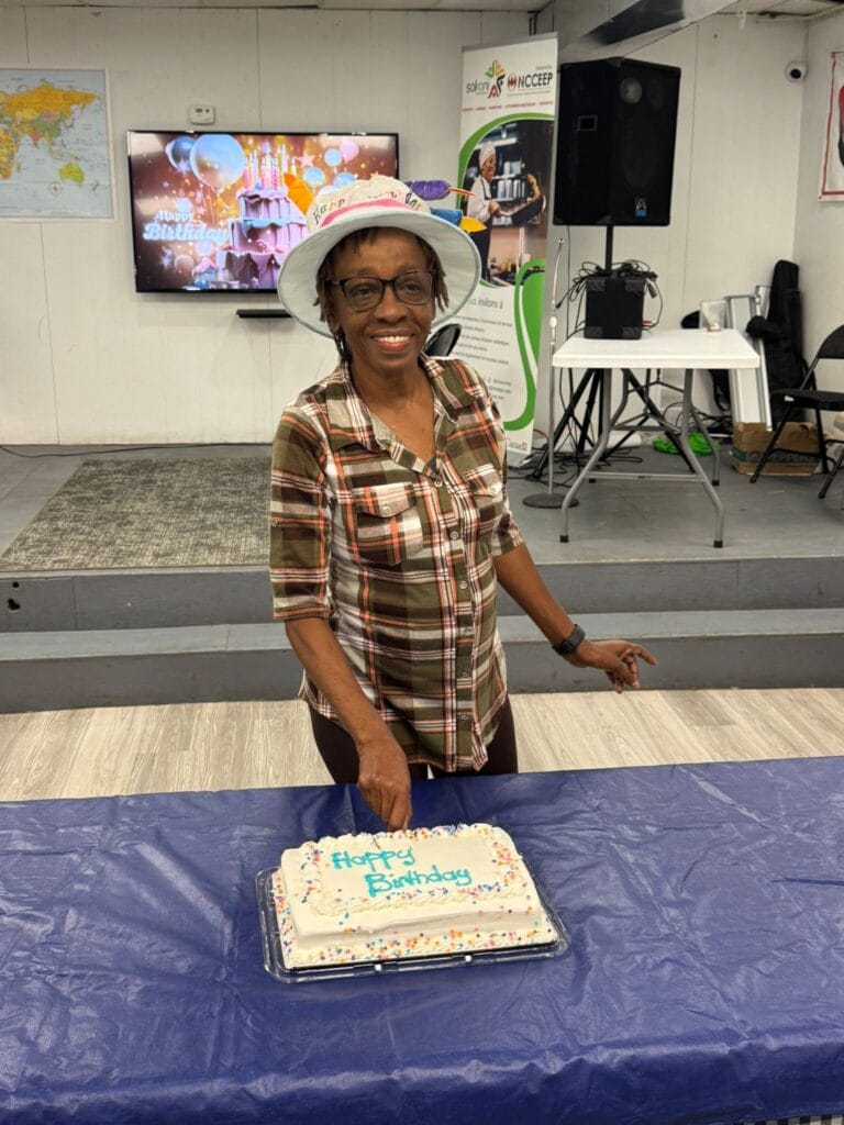 A woman in a checked shirt and a paper party hat smiling behind the cake with a knife in her hand